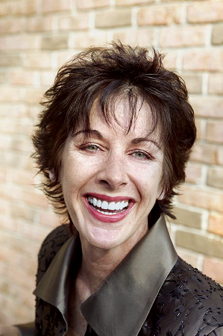 The image shows a woman with short hair, smiling broadly, wearing a dark top and sitting against a brick wall.