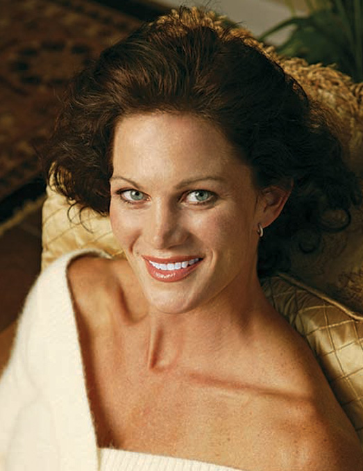 A woman with long hair, wearing a white top, sitting and smiling at the camera.