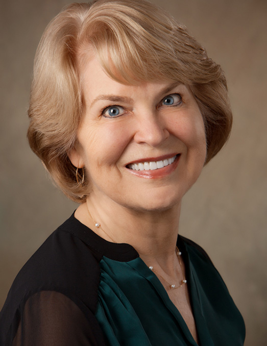 The image depicts a woman with short blonde hair, wearing a dark top and a necklace, smiling at the camera against a neutral background.