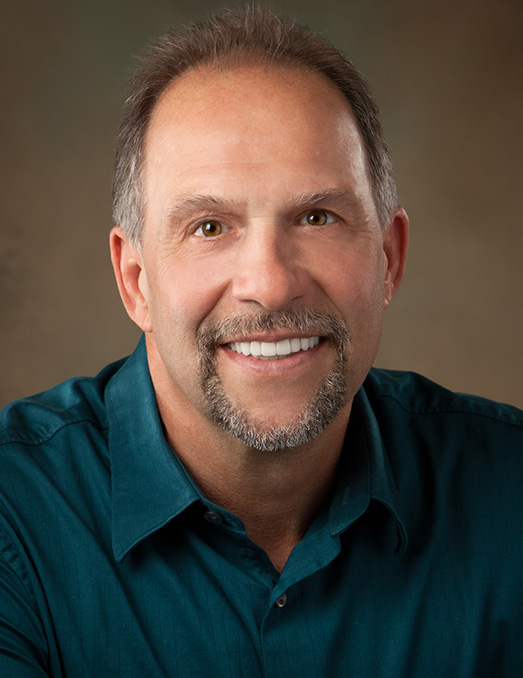 The image is a color photograph of a man posing for a portrait. He has a beard and mustache, is smiling, and appears to be in his middle years. The man is wearing a dark blue shirt with the sleeves rolled up and has short gray hair. His expression is friendly and confident.