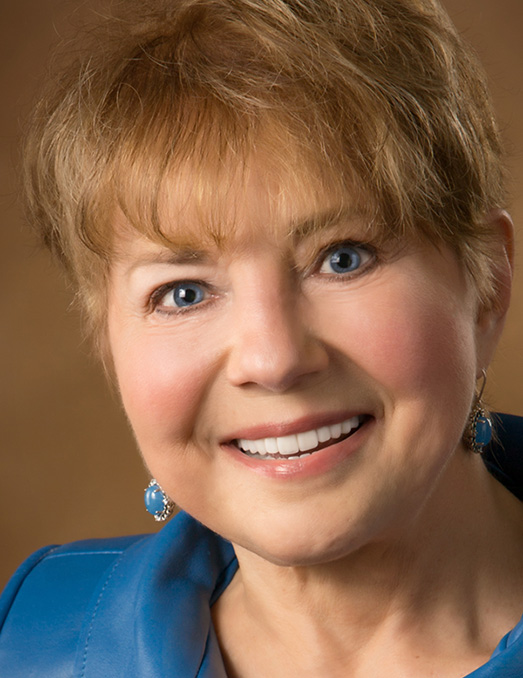 A smiling woman with short hair, wearing a blue jacket and a necklace, posing for a portrait.