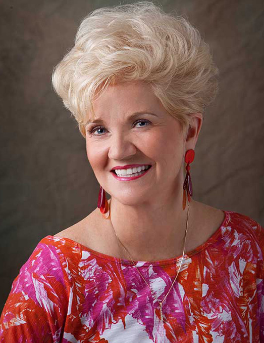 A woman with blonde hair, wearing a pink top and earrings, smiling at the camera.