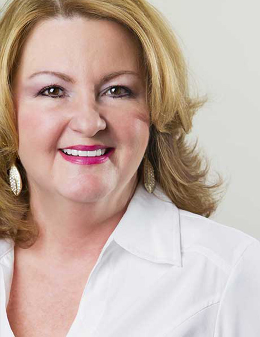The image shows a woman with short brown hair, wearing a white top and a dark blazer. She has a confident expression and is looking directly at the camera.