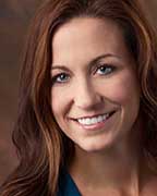 A smiling woman with long brown hair, wearing a blue top and a necklace, posing for a portrait.