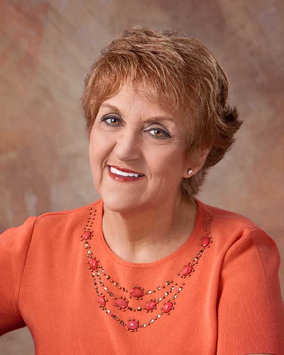 The image features a woman with short hair, wearing an orange top and a necklace, posing for the camera with a slight smile.