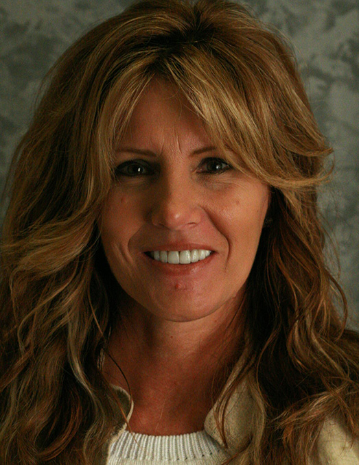 The image is a photograph of a woman with long blonde hair, wearing a light-colored top and a necklace. She has a friendly expression and is smiling at the camera.