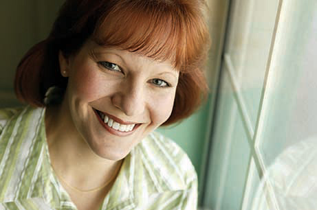 The image is a photograph of a woman with red hair, wearing a light-colored top and a smile.