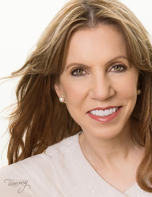 Woman with smiling expression, wearing a white top and earrings, against a blurred background.