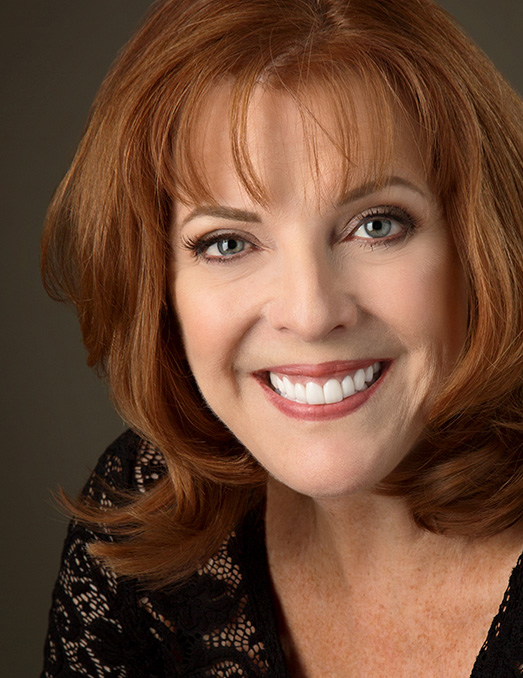 The image features a woman with red hair, wearing a black top and a necklace, smiling at the camera against a dark background.