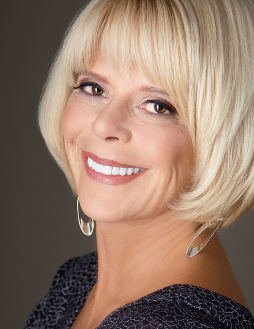 Woman with short blonde hair, smiling at the camera, wearing a dark top and a necklace.
