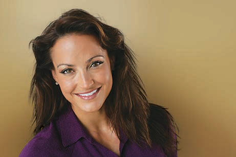 A woman with long hair is smiling at the camera, wearing a purple top.