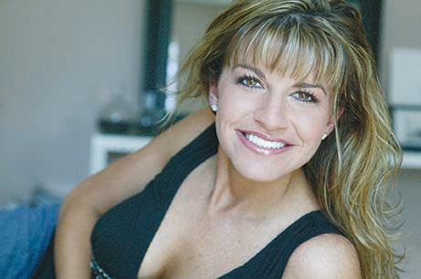 The image shows a woman posing for the camera with a smile, wearing a sleeveless top and leaning slightly to one side.