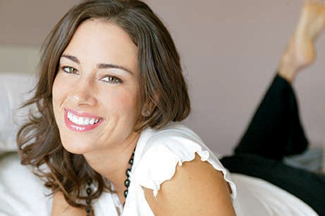 A woman with a radiant smile, lying down on her stomach with one hand resting on her hip, against a plain background.