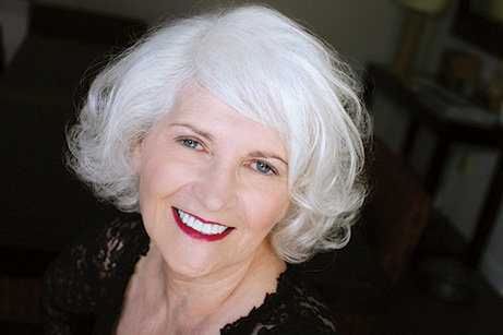 An elderly woman with white hair, wearing a black top and red lipstick, poses for the camera.