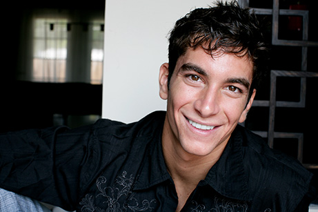 The image is a portrait of a man with a beard, smiling at the camera. He has short hair and is wearing a dark shirt. There s a warm and welcoming vibe to his expression.