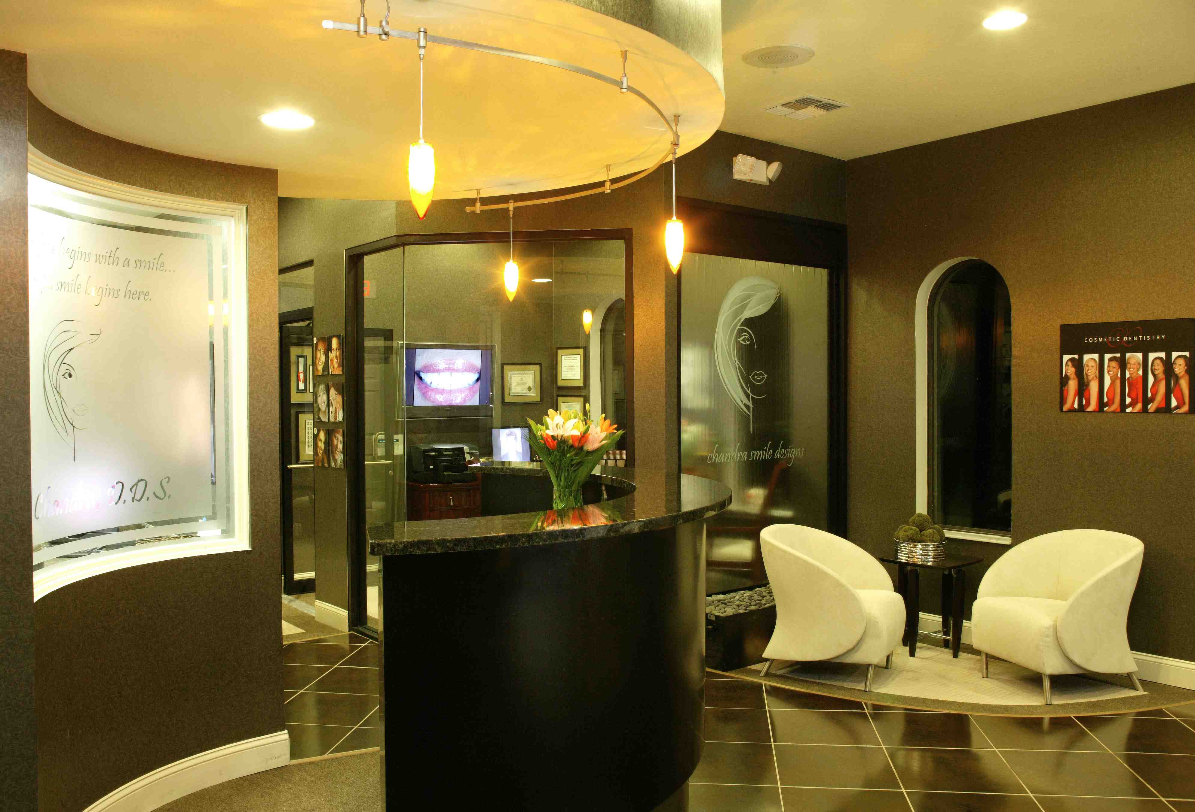 The image depicts an interior space, possibly a lobby or waiting area of a dental office or salon. There is a modern design with a neutral color palette, featuring a reception desk, chairs, and decorative elements such as framed certificates on the walls.