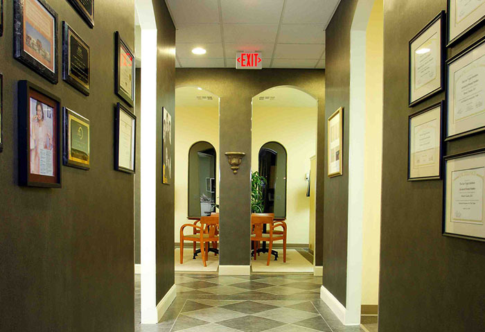 The image depicts an interior hallway with a framed certificate on the wall, a door at the end, and a series of framed items lining the wall.