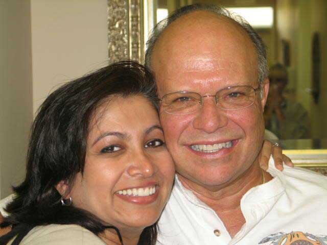 The image is a photograph of an older man and a younger woman posing closely together, with the man smiling broadly and the woman looking at the camera with a smile.