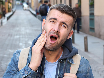 The image features a man with a beard and mustache, wearing a denim jacket and holding his hand to his mouth as if he s in the middle of speaking or singing. He appears surprised or reacting to something off-camera. The background suggests an urban street scene with buildings and pedestrians.