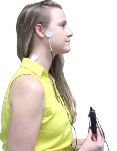 A woman stands with a device attached to her ear, likely for hearing assistance or testing.