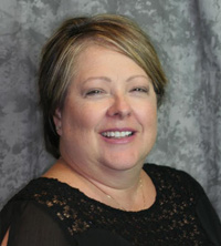 A smiling woman in a black dress, posing for a portrait with a grey background.