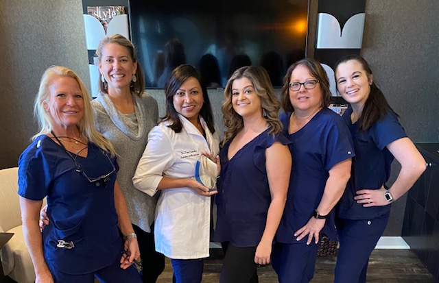 The image shows a group of six individuals, likely healthcare professionals, posing together in front of a television screen. They are dressed in scrubs and standing in front of a wall with a framed diploma or certificate.