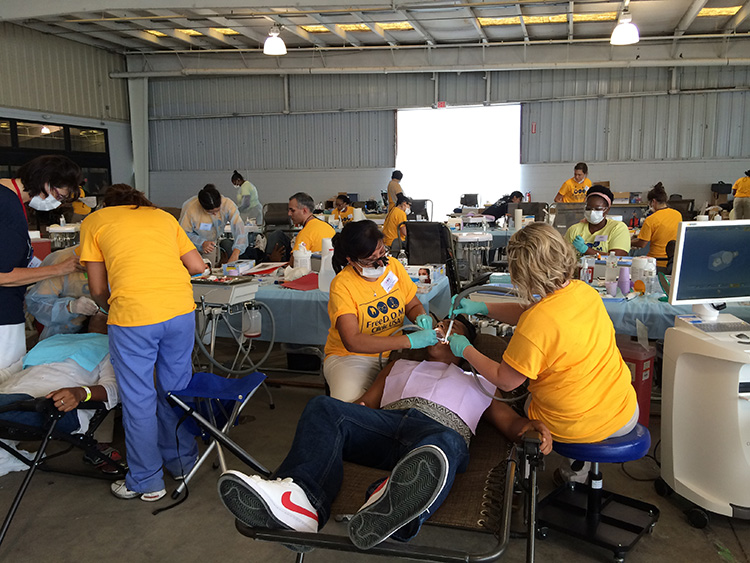 The image shows a room with multiple people, some of whom are seated in dental chairs. There is medical equipment visible and the setting appears to be a makeshift dental or medical care facility.