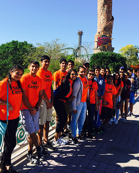 A group of people, likely a team or school group, posing for a photo in front of a theme park attraction with their shirts displaying various messages and logos.