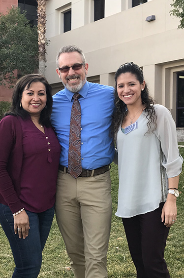 The image shows three individuals posing together outdoors, with a man in the center wearing a blue shirt and tie, flanked by two women  one is wearing a maroon top and the other has a light-colored blouse. They are all smiling at the camera.