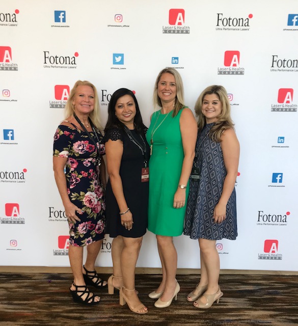 A group of four women posing for a photo at an event, standing in front of a backdrop with logos and text.
