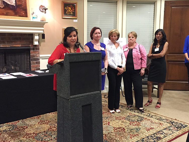 The image shows a woman standing at a podium, giving a speech or presentation, with an audience of people standing behind her.