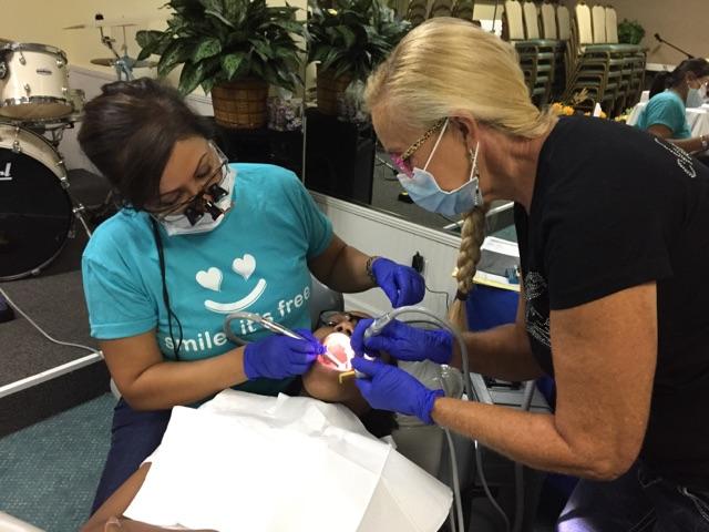 Two women wearing face masks, one is performing a dental procedure on the other while seated in chairs.