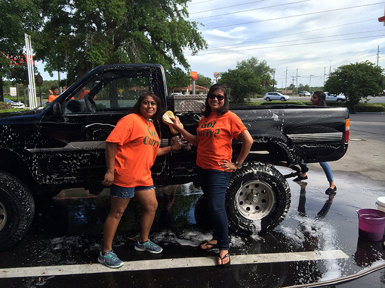 Two women posing in front of a black pickup truck parked at a car wash, with one woman holding a mop bucket and the other smiling.