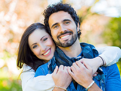 A man and a woman are embracing each other in a warm hug, smiling and looking directly at the camera.