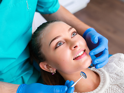 A dental professional is adjusting a woman s teeth while she smiles, with both of them wearing blue gloves and the setting appears to be a dental office.