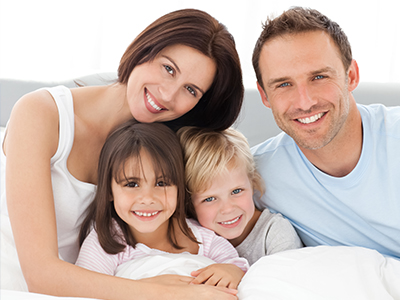 A family of four posing for a photograph in bed, with the father smiling at the camera and the mother holding the child.