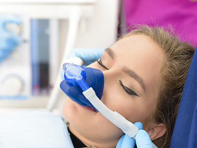 A person receiving medical care, with a blue oxygen mask over their mouth and a clear plastic tube attached to it, being attended to by a healthcare professional wearing gloves.