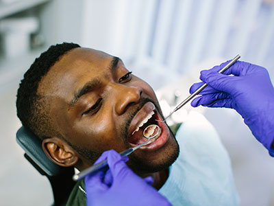 The image shows a person in a dental chair receiving dental care, with a dentist using instruments to work on their teeth while the patient is wearing protective eyewear.